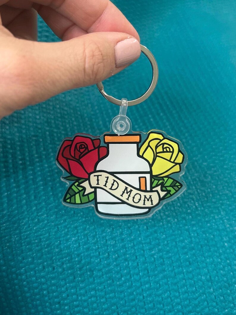 Op de afbeelding: Een transparante acryl sleutelhanger met een cartoonillustratie van een pillenfles met de tekst "TID MOM" op een banner. De fles wordt omringd door twee rozen, een rode en een gele.