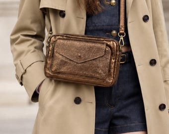 May include: A rectangular metallic bronze crossbody bag with a front flap pocket and a thin adjustable leather strap. The bag features a textured finish and gold-tone hardware, worn over a dark denim outfit and a beige trench coat.