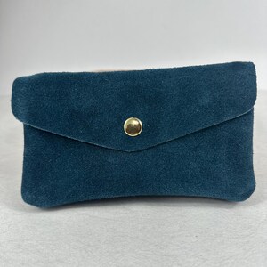 Op de afbeelding: Teal suède portemonnee met een klepsluiting en een goudkleurige drukknop. De rechthoekige portemonnee heeft een zachte, getextureerde uitstraling. Het ontwerp is eenvoudig en elegant, geschikt voor het dragen van kaarten of kleine items.