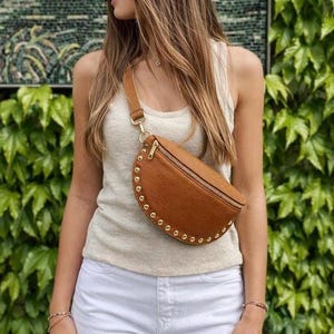 Peut inclure: Un sac banane en cuir marron avec une fermeture éclair dorée et des clous dorés. Le sac est porté en bandoulière. La sangle est réglable. La personne porte un débardeur blanc et un jean blanc.