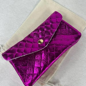 May include: A metallic fuchsia clutch bag with a quilted pattern and a gold-coloured snap closure. The clutch is open, revealing a beige fabric pouch inside. The clutch is rectangular with a flap closure.