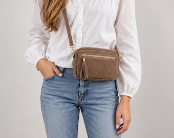 May include: A brown rectangular crossbody bag with a textured finish, featuring a front zippered pocket with a matching tassel pull, a gold-tone metal zipper, and a thin adjustable shoulder strap worn across the torso.