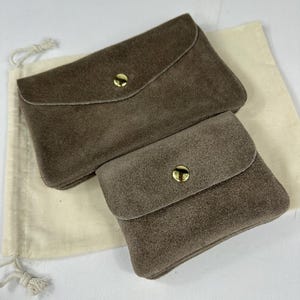 Puede incluir: Dos bolsos de ante color topo con solapas redondeadas estilo sobre y cierres de presión de metal dorado. Los bolsos tienen forma rectangular y presentan una superficie suave y texturizada. Están colocados sobre una bolsa de tela color crema.