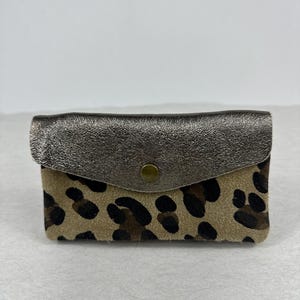 Puede incluir: Una pequeña cartera rectangular con cierre de solapa. La solapa es de color plateado metálico y el cuerpo de la cartera presenta un fondo beige con manchas de leopardo marrón oscuro. Un pequeño broche dorado asegura la solapa.