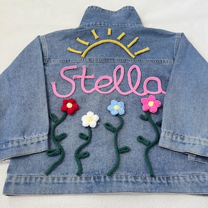 May include: A blue denim jacket with a pink embroidered name "Stella" and a yellow embroidered sun above it.  There are four crocheted flowers in red, white, blue, and pink with green crocheted stems below the name.