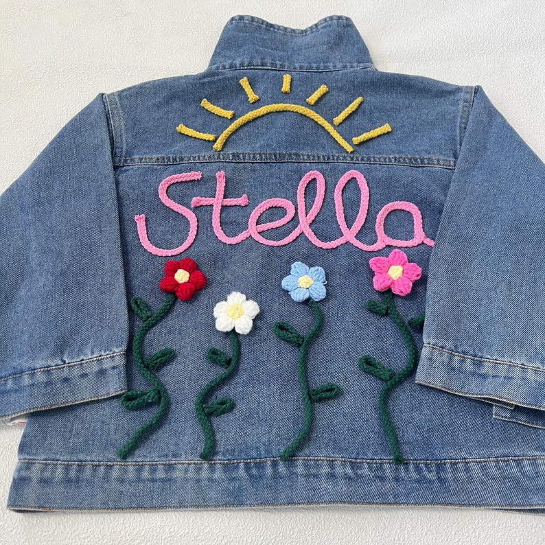 May include: A blue denim jacket with a pink embroidered name "Stella" and a yellow embroidered sun above it.  There are four crocheted flowers in red, white, blue, and pink with green crocheted stems below the name.