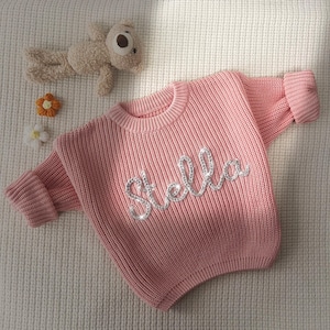Könnte beinhalten: Ein rosa gestrickter Pullover mit dem Namen "Stella" in silbernen Glitzer.