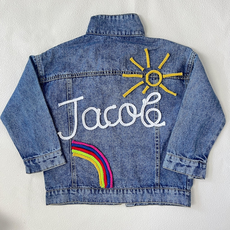 May include: A blue denim jacket with white embroidery of the name "Jacob" and a yellow sun with a rainbow below it.