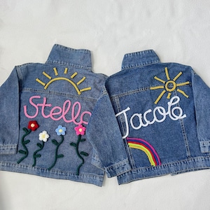 May include: Two denim jackets with personalized embroidery. One jacket features a pink "Stella" with flowers and a yellow sun. The other jacket features a white "Jacob" with a rainbow and a yellow sun.