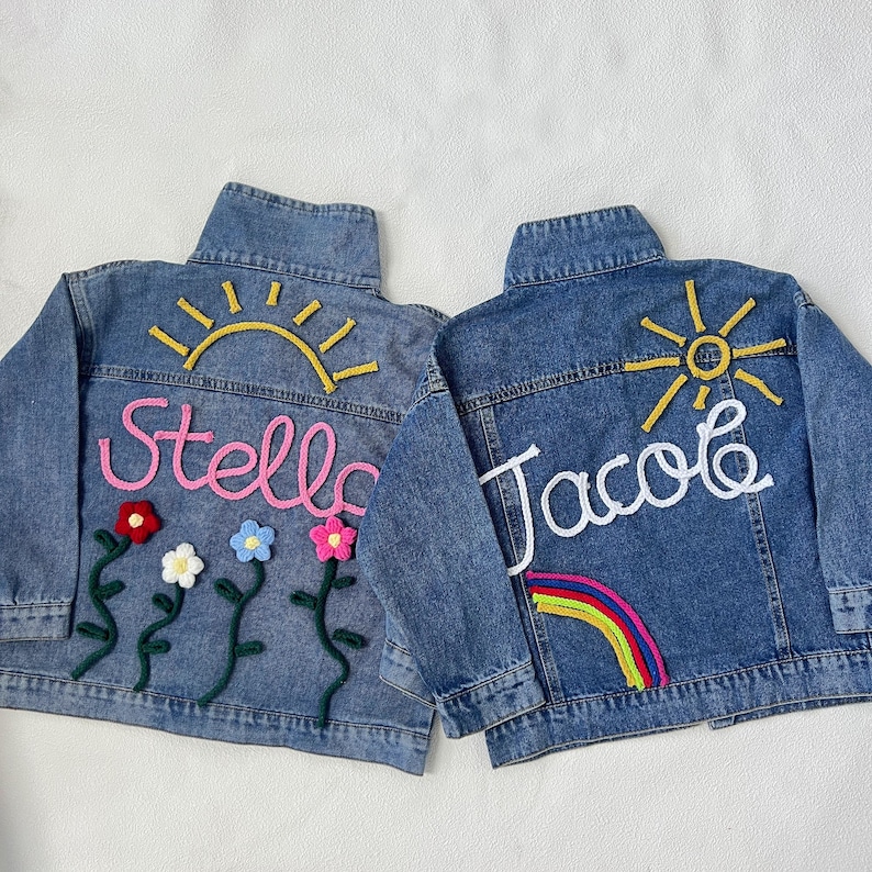 May include: Two denim jackets with personalized embroidery. One jacket features a pink "Stella" with flowers and a yellow sun. The other jacket features a white "Jacob" with a rainbow and a yellow sun.