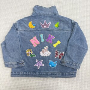 May include: A blue denim jacket with various colorful patches, including a crown, a star, a mermaid, a butterfly, a castle, a whale, a starfish, and an avocado. The name "NIKI" is spelled out in sparkly letters.