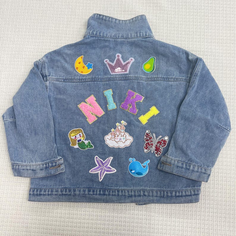 May include: A blue denim jacket with various colorful patches, including a crown, a star, a mermaid, a butterfly, a castle, a whale, a starfish, and an avocado. The name "NIKI" is spelled out in sparkly letters.