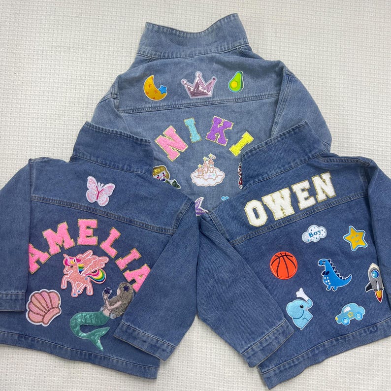May include: Three denim jackets with colorful patches and embroidered names. The jackets have a variety of patches, including a unicorn, a mermaid, a dinosaur, a basketball, a star, and a crown. The names on the jackets are Amelia, Owen, and Niki.
