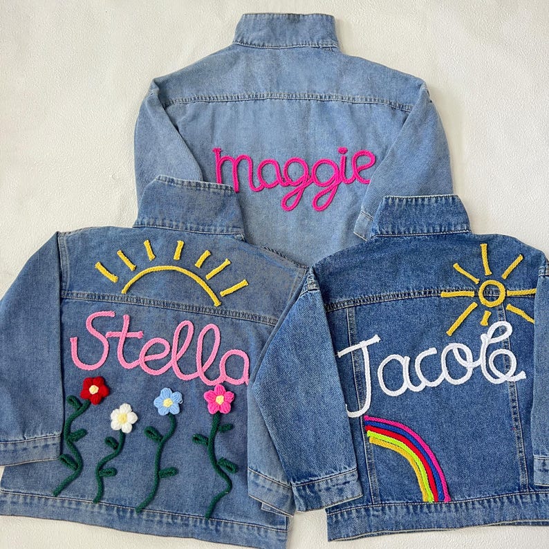 May include: Three denim jackets with embroidered names and designs. The jackets are blue and feature the names "Maggie", "Stella", and "Jacob" in colorful yarn. Stella's jacket has a sun and flowers, Jacob's has a sun and rainbow.