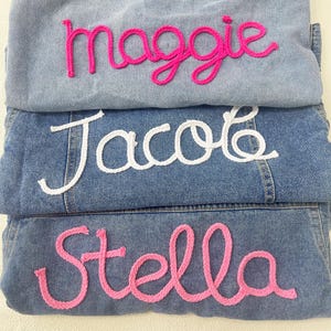 May include: Three denim jackets with names embroidered on them in pink and white yarn. The names are "Maggie", "Jacob", and "Stella".