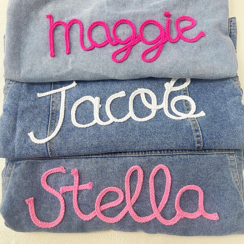 May include: Three denim jackets with names embroidered on them in pink and white yarn. The names are "Maggie", "Jacob", and "Stella".