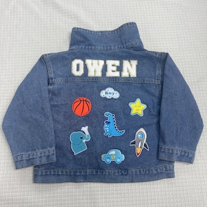 May include: A blue denim jacket with the name "OWEN" embroidered on the back. The jacket has several colorful patches sewn on, including a basketball, a dinosaur, a star, a rocket, a car, and an elephant.