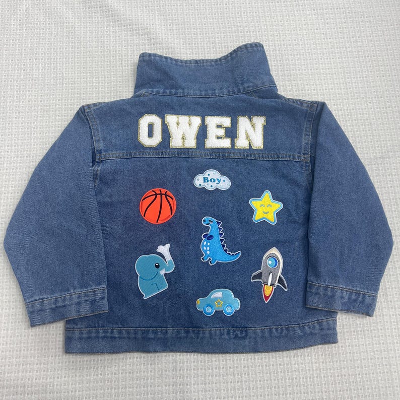 May include: A blue denim jacket with the name "OWEN" embroidered on the back. The jacket has several colorful patches sewn on, including a basketball, a dinosaur, a star, a rocket, a car, and an elephant.