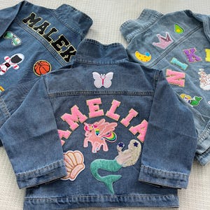 May include: Three denim jackets with colorful patches and embroidered names. The jackets are blue denim with a variety of patches including a unicorn, mermaid, butterfly, crown, avocado, and stars. The names on the jackets are "Malek", "Amelia", and "Kim".