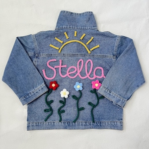 May include: A blue denim jacket with a yellow sun, pink yarn lettering that says "Stella", and green yarn flowers with pink, blue, white, and red centers.