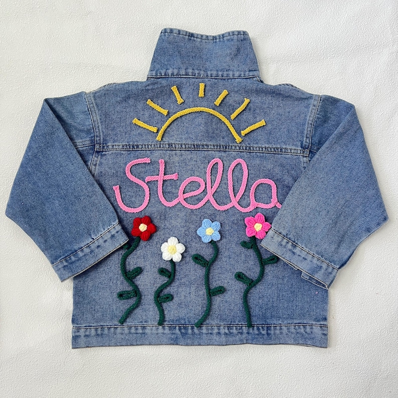 May include: A blue denim jacket with a yellow sun, pink yarn lettering that says "Stella", and green yarn flowers with pink, blue, white, and red centers.