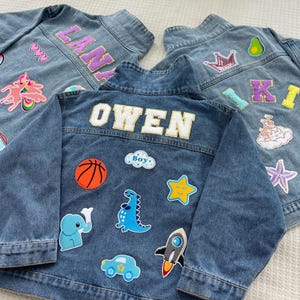 May include: Two denim jackets with colorful patches and embroidered names. One jacket has the name "OWEN" and patches of a basketball, dinosaur, rocket, car, elephant, and a cloud with the word "Boy". The other jacket has the name "KIKI" and patches of a crown, avocado, castle, and a starfish.