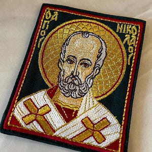 Puede incluir: Parche bordado que representa una figura religiosa con aureola y barba doradas. El parche tiene un fondo verde oscuro con detalles rojos y dorados. La figura lleva una vestimenta blanca y dorada con cruces doradas.