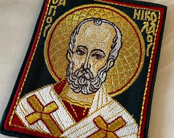 Könnte beinhalten: Gestickter Aufnäher mit einer religiösen Figur mit goldenem Heiligenschein und Bart. Der Aufnäher hat einen dunkelgrünen Hintergrund mit roten und goldenen Akzenten. Die Figur trägt ein weißes und goldenes Gewand mit goldenen Kreuzen.