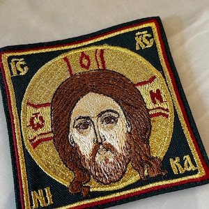 Może przedstawiać: Haftowana naszywka przedstawiająca Jezusa Chrystusa z aureolą. Projekt zawiera złote, brązowe i czerwone nici na ciemnozielonym tle, z tekstem religijnym. Naszywka ma kształt kwadratu.