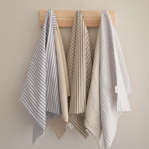 May include: Three striped kitchen towels in blue and white, beige, and gray and white hang from a wooden rack. The towels are made of a soft fabric and have a classic, farmhouse style.
