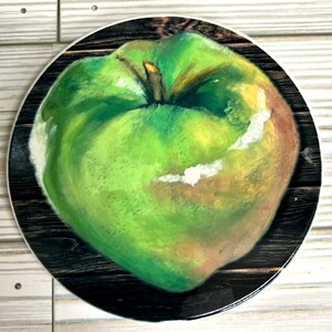 May include: A green apple with a brown stem and a white spot on the side, painted on a black and brown wooden background.