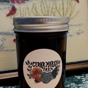 Könnte beinhalten: Ein Glasgefäß mit schwarzem Deckel und einem weißen Etikett mit der Aufschrift "Cedar Marsh Farm". Das Etikett zeigt eine Zeichnung eines Pilzes, eines Kohlkopfes und einer Himbeere.