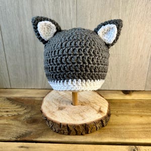 May include: Grey and white crocheted hat with wolf ears. The hat is on a wooden stand.