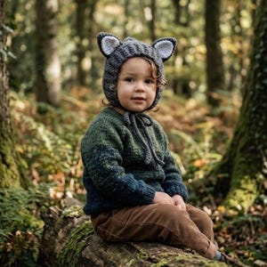 May include: A child wearing a gray knit hat with white-lined ears and a green and blue knit sweater. The child is also wearing brown corduroy pants. The child is sitting on a moss-covered log in a forest setting.