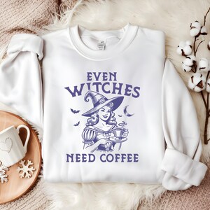 Op de afbeelding: Witte sweatshirt met de paarse tekst "EVEN WITCHES NEED COFFEE". De afbeelding toont een vintage-stijl illustratie van een heks die een kopje koffie vasthoudt, met vleermuizen en een maan.