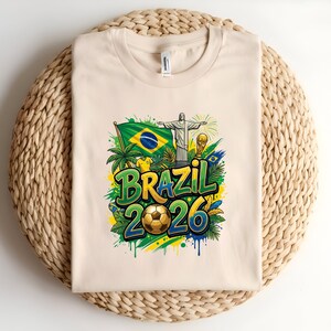 Cream t-shirt with Brazil 2026 graphic including flag, statue, and soccer ball.