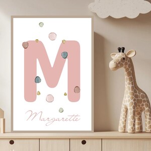 Pode incluir: Uma impressão emoldurada com uma grande letra "M" rosa e o nome "Margarette" em escrita cursiva. A letra é adornada com pequenos pontos de aquarela coloridos. A impressão é exibida em um armário de madeira clara, com uma estatueta de girafa e outros itens decorativos.