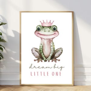 Pode incluir: Uma ilustração em aquarela de um sapo verde usando uma coroa rosa. O sapo está sentado em um fundo branco com o texto "dream big little one" abaixo.