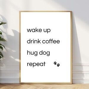 Könnte beinhalten: Ein gerahmter Druck mit weißem Hintergrund und schwarzem Text mit der Aufschrift "wake up, drink coffee, hug dog, repeat". Zwei schwarze Pfotenabdrücke befinden sich unten. Der Rahmen ist hellbraun.