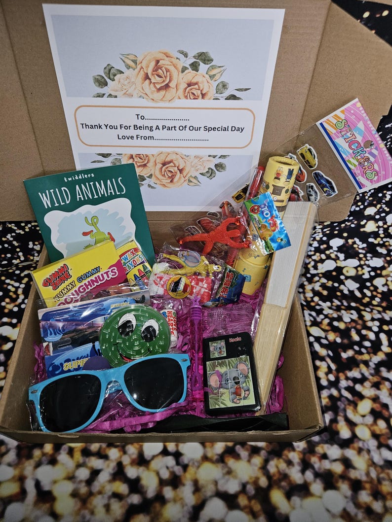 May include: A brown cardboard box filled with a variety of children's toys and treats. The box includes a book titled "Wild Animals", a pair of blue sunglasses, a small wooden toy, a bag of gummy candies, a bag of wax crayons, a bag of stickers, and a small plastic toy. The box is decorated with a card that says "Thank You For Being A Part Of Our Special Day".