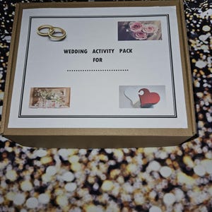 May include: A brown cardboard box with a white label that says "Wedding Activity Pack For". The label also has images of two gold rings, a bouquet of pink roses, and two wooden hearts.