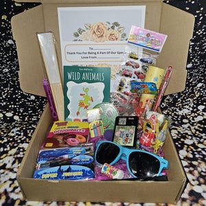 May include: A brown cardboard box filled with various items, including a book titled "Wild Animals", a pack of stickers, a pack of crayons, a pair of sunglasses, and a bag of gummy donuts. The box also includes a card with the text "Thank You For Being A Part Of Our Special Love From..."