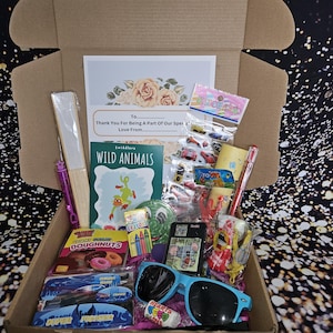 May include: A brown cardboard box filled with various items, including a coloring book titled "Wild Animals", crayons, stickers, sunglasses, a fan, and a bag of donuts. The box also includes a card with the text "To... Thank You For Being A Part Of Our Special... Love From..."