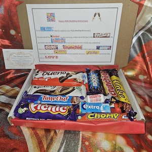 May include: A gift box filled with a variety of chocolate bars, including Galaxy, Time Out, Crunchie, Flake, Picnic, Bueno, Boost, and Extra. The box is decorated with red and white paper and a card that says "Happy 40th Wedding Anniversary".