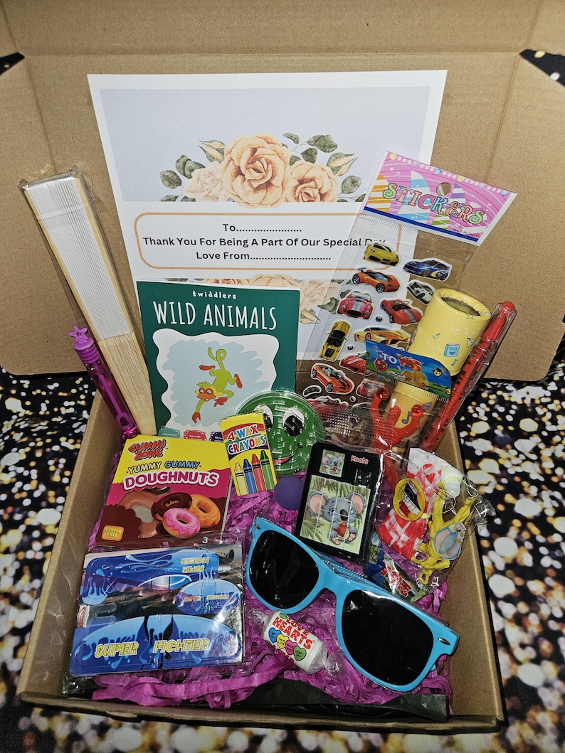May include: A gift box filled with various items, including a coloring book, crayons, stickers, a fan, sunglasses, and candy. The box is lined with pink tissue paper. The box has a card with the text "Thank You For Being A Part Of Our Special Day Love From..."