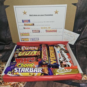 May include: A cardboard box filled with a variety of chocolate bars, including Crunchie, Yorkie, Kinder Bueno, Wispa, Picnic, Lion, and Starbar. The box is decorated with a red ribbon and a card that says "Well done on your Promotion".