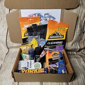 May include: A brown cardboard box containing car cleaning products, including Armor All wipes and a demist pad. Other items include a PG Tips tea bag, a Yorkie chocolate bar, and small bottles of Lynx and Nescafé.