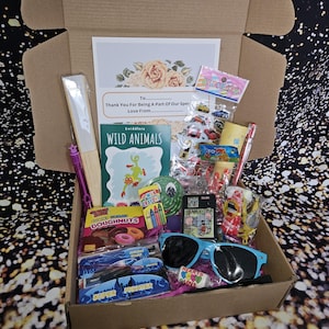 May include: A brown cardboard box filled with various items, including a book titled "Wild Animals", a bag of gummy doughnuts, a pair of blue sunglasses, and a variety of colorful toys and trinkets. The box is open and the items are visible inside.