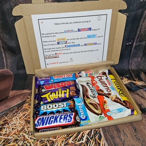May include: A brown cardboard box filled with a variety of chocolate bars, including Twirl, Wispa, Galaxy, Kinder Bueno, and Snickers. The box is decorated with a birthday message and a list of chocolate brands.