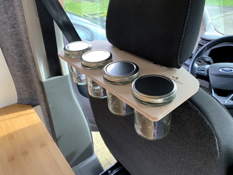 May include: A wooden shelf with four glass jars with silver lids attached to the headrest of a car seat. The shelf is labeled "Camp Kitchen".